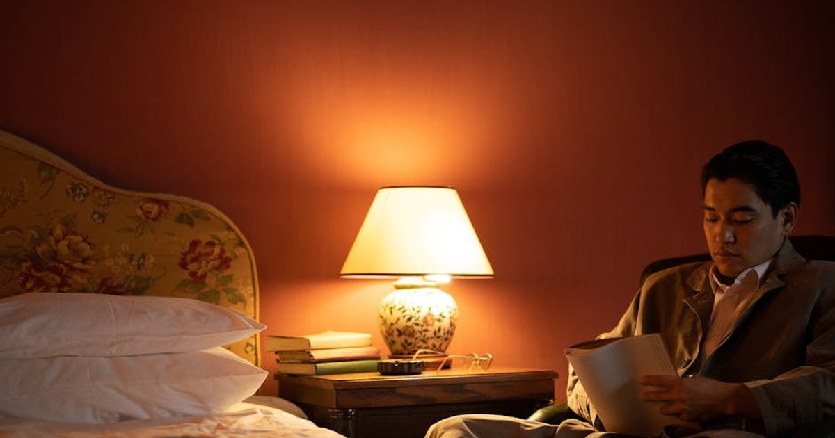 A man sits in a cozy hotel room reading under warm, ambient light.