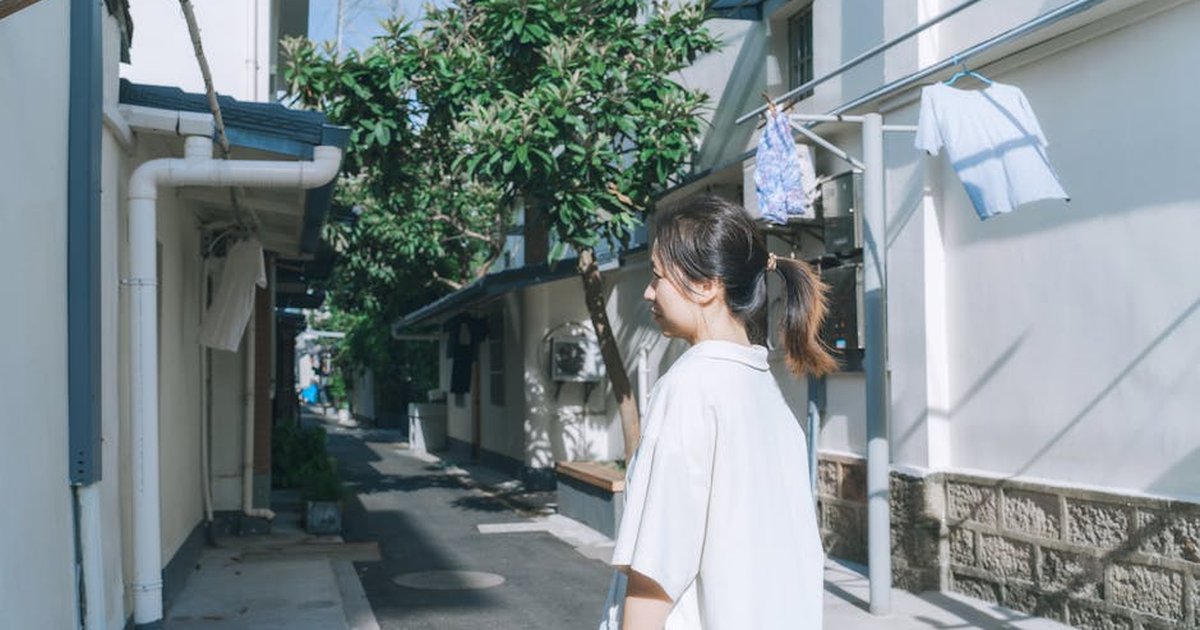 A young woman walks along a sunlit alleyway with hanging laundry and green foliage.