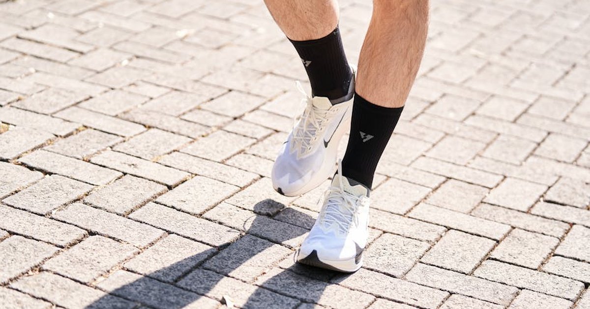 A person wearing white sneakers and black socks walking on a sunlit brick pathway.
