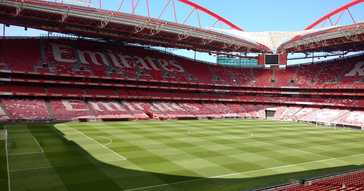 A large empty soccer stadium with red seats and a well-maintained green field under a clear blue sky.