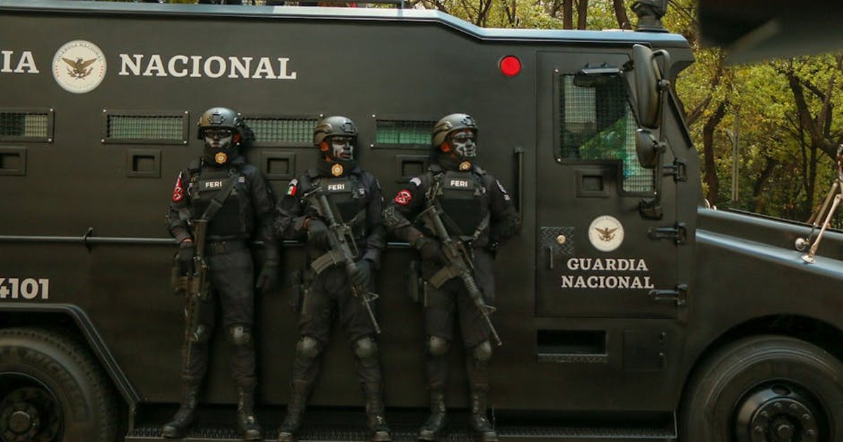 Guardia Nacional officers in tactical gear stand alert beside an armored vehicle.