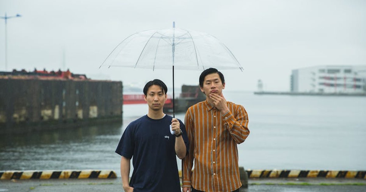 Serious young Asian males in casual wear with umbrella in rainy weather on embankment of river
