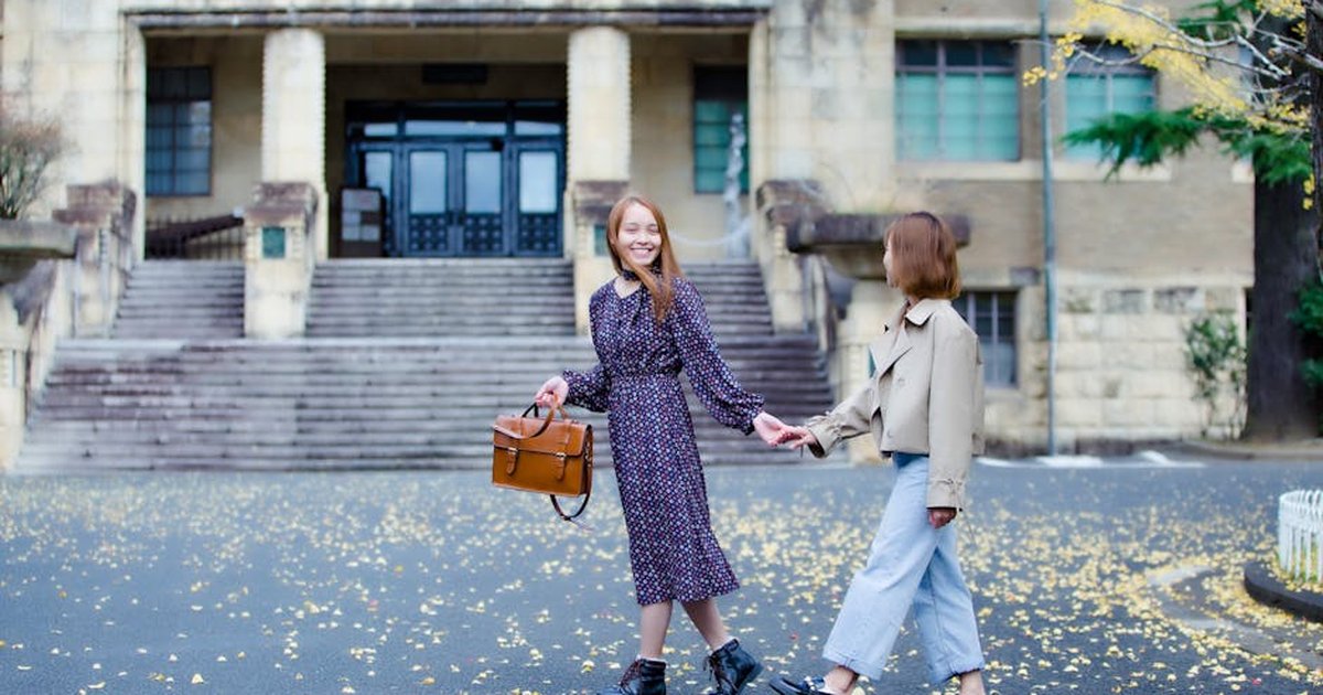 Two women holding hands and smiling while walking past a historic building in an autumn setting.