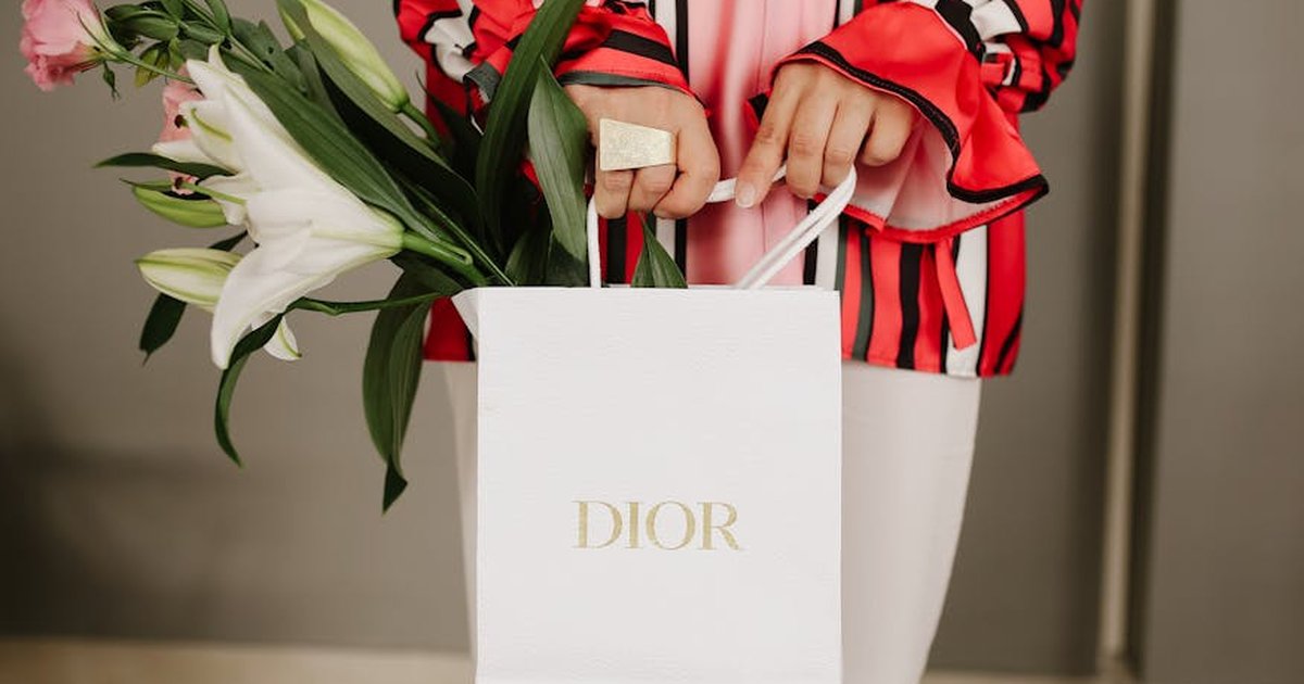 Close-up of a woman holding a Dior paper bag with lilies and roses, wearing a stylish outfit indoors.