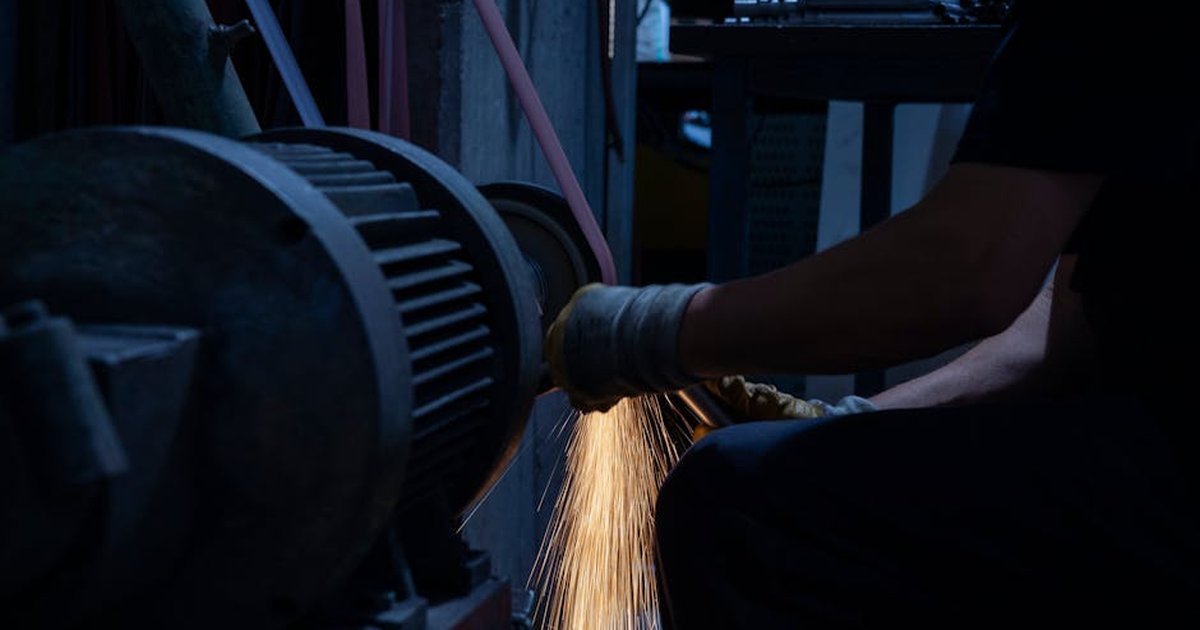 A worker in a factory uses a grinding machine, generating bright sparks in a dark industrial setting.