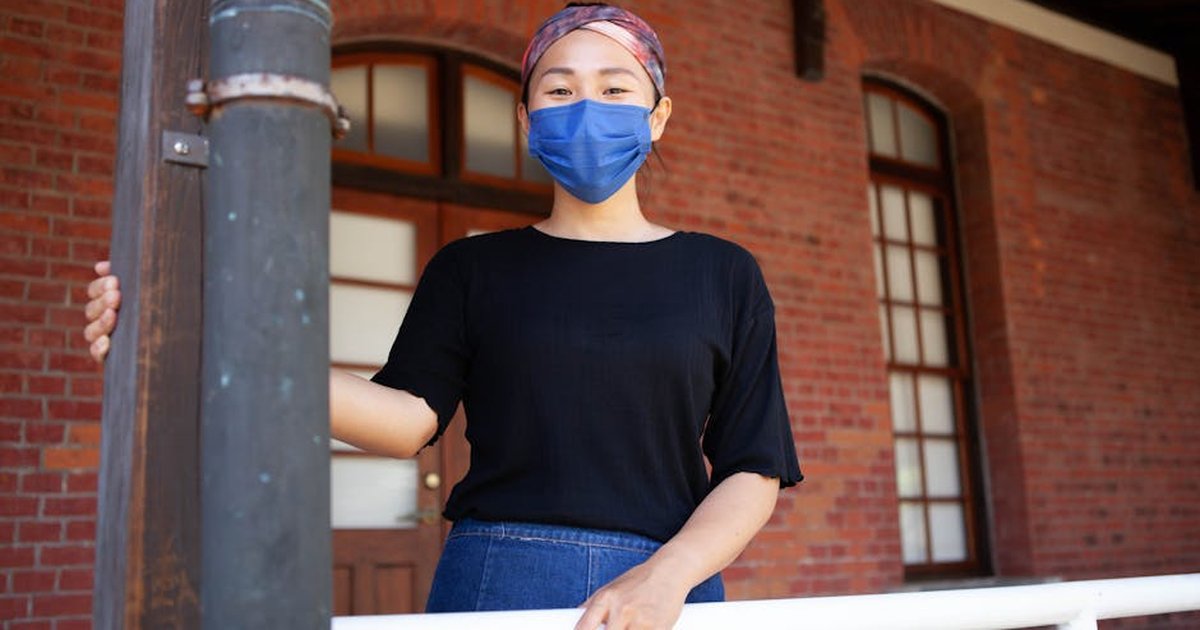 A young woman with a face mask stands outdoor against a red brick wall, embodying new normal life.