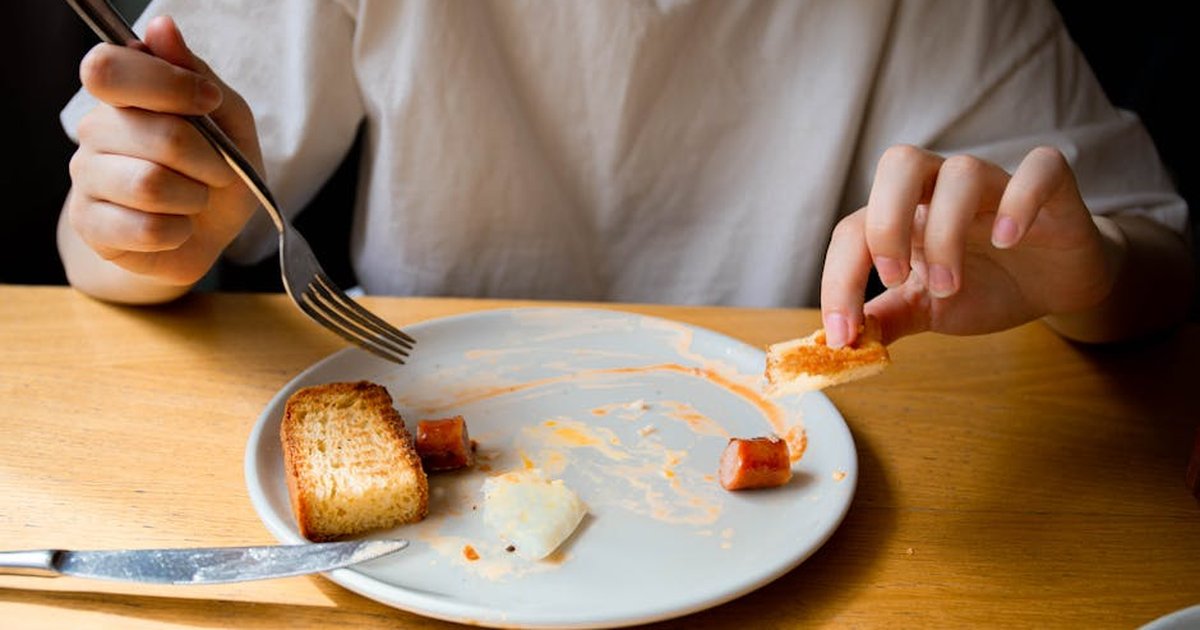 A person enjoying a casual meal of toasted bread and sausage on a white plate at a wooden table.