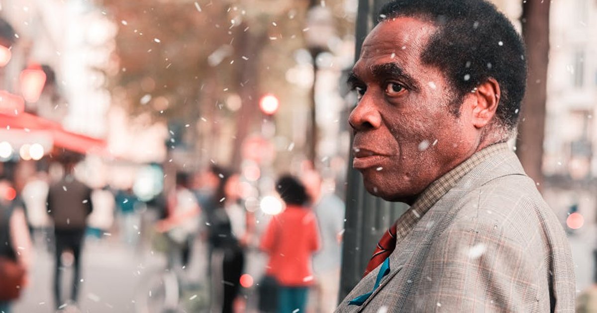 A man stands amidst a bustling city street under a gentle snowfall, captured in warm tones.