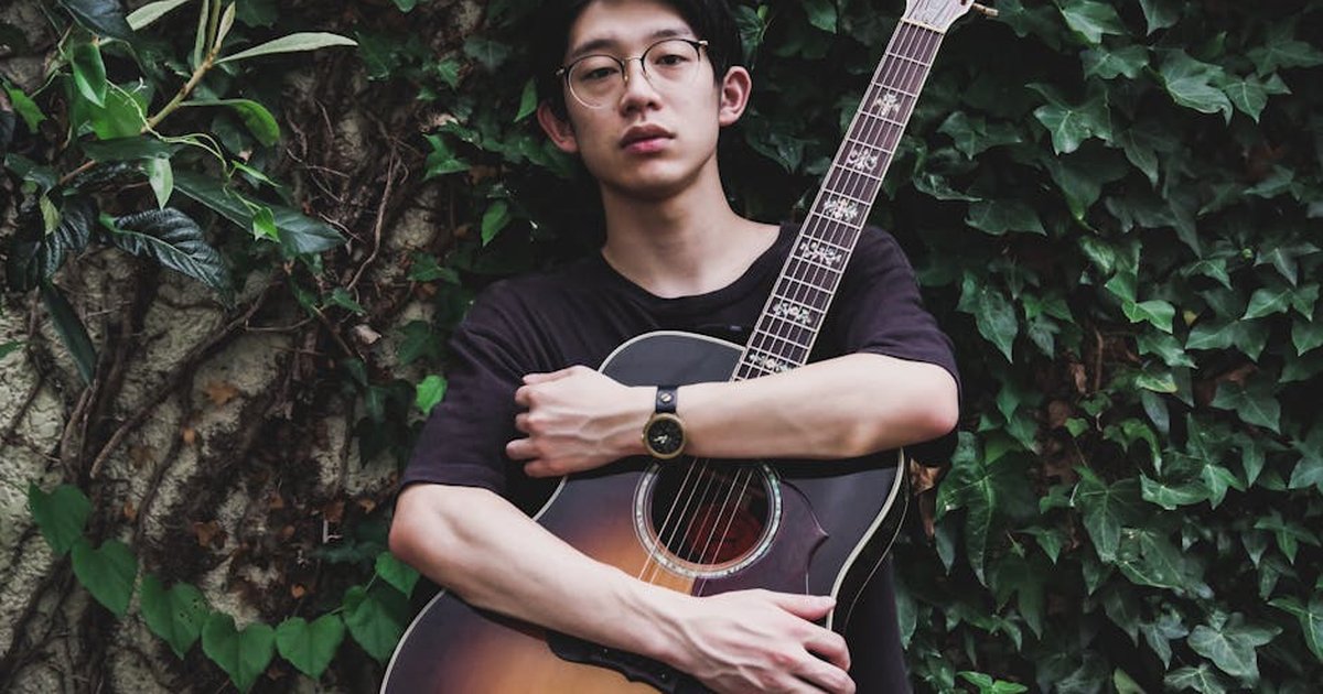 Young Asian man with glasses holding an acoustic guitar in front of green ivy wall.