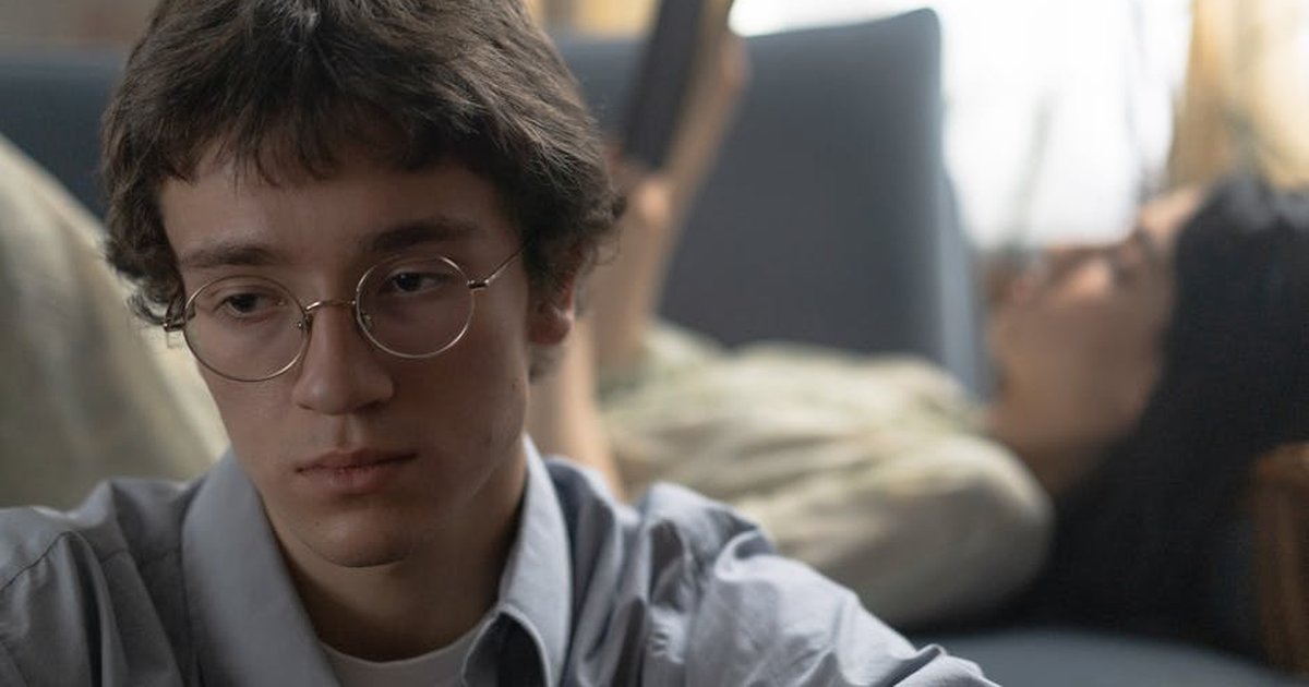 Young man in glasses looks thoughtful while a woman reads in the background, indoors.