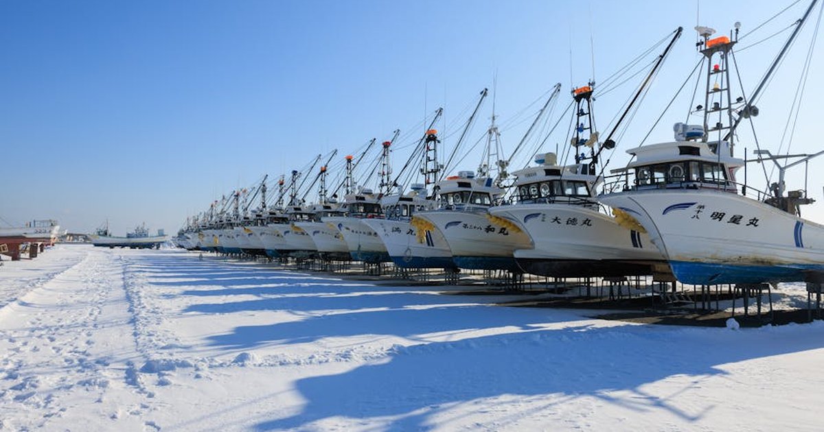 Fishing boats lined up on a snowy harbor in Hokkaido, Japan, under a clear blue sky.