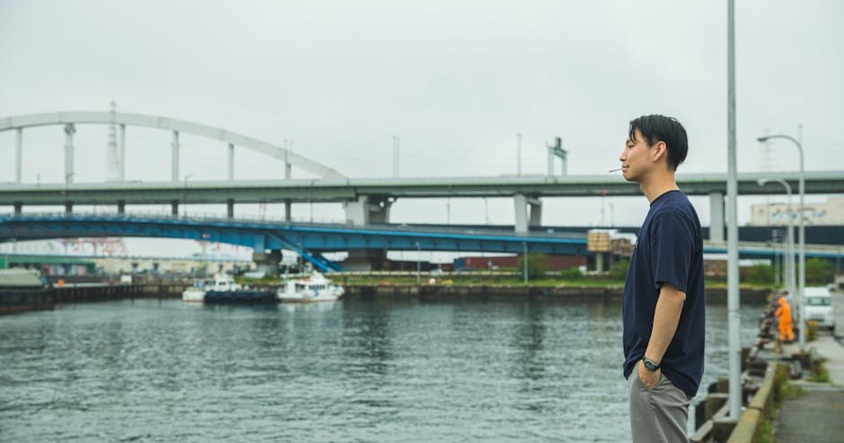 Side view of Asian young man in casual outfit with cigarette and hands in pockets standing on embankment and thinking