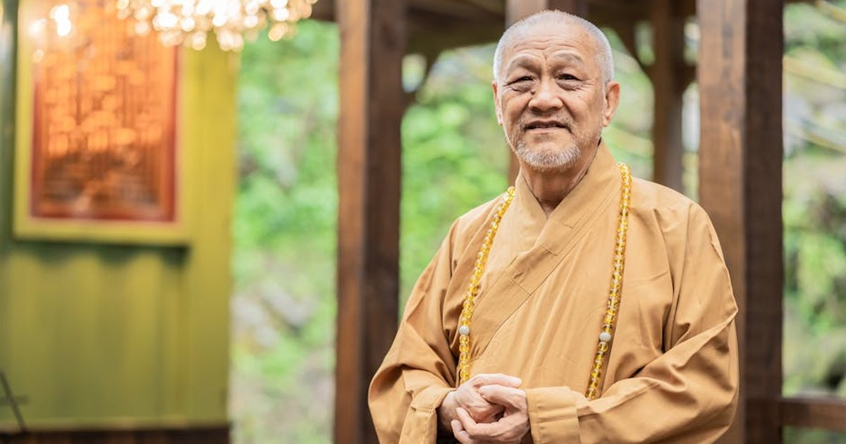 Elderly Buddhist monk standing indoors in traditional garments. Spiritual and peaceful setting.