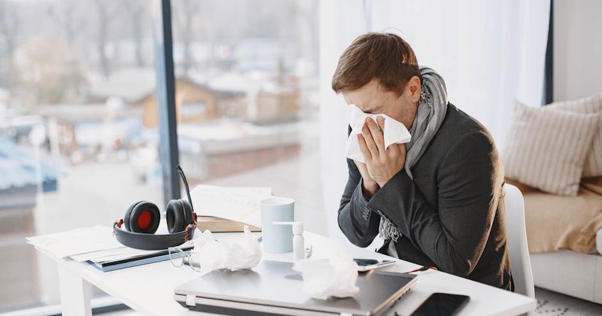 Man sneezing at home desk, indicating remote work during illness.