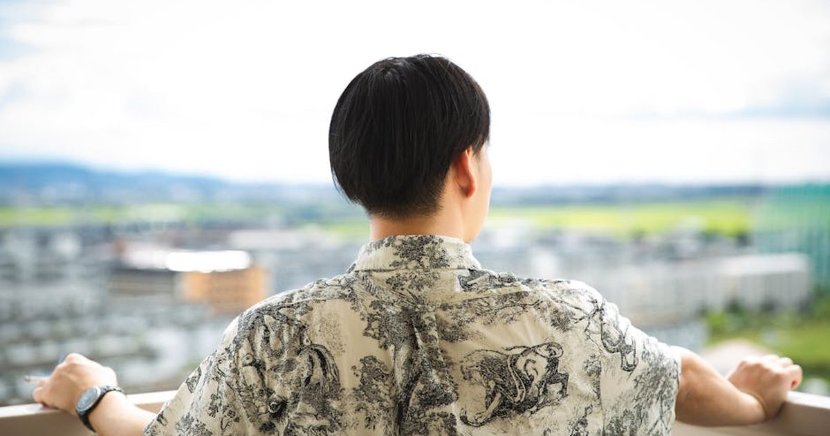 Back view anonymous brunet in trendy shirt standing on open balcony and enjoying view of small town located in hilly terrain on sunny summer day