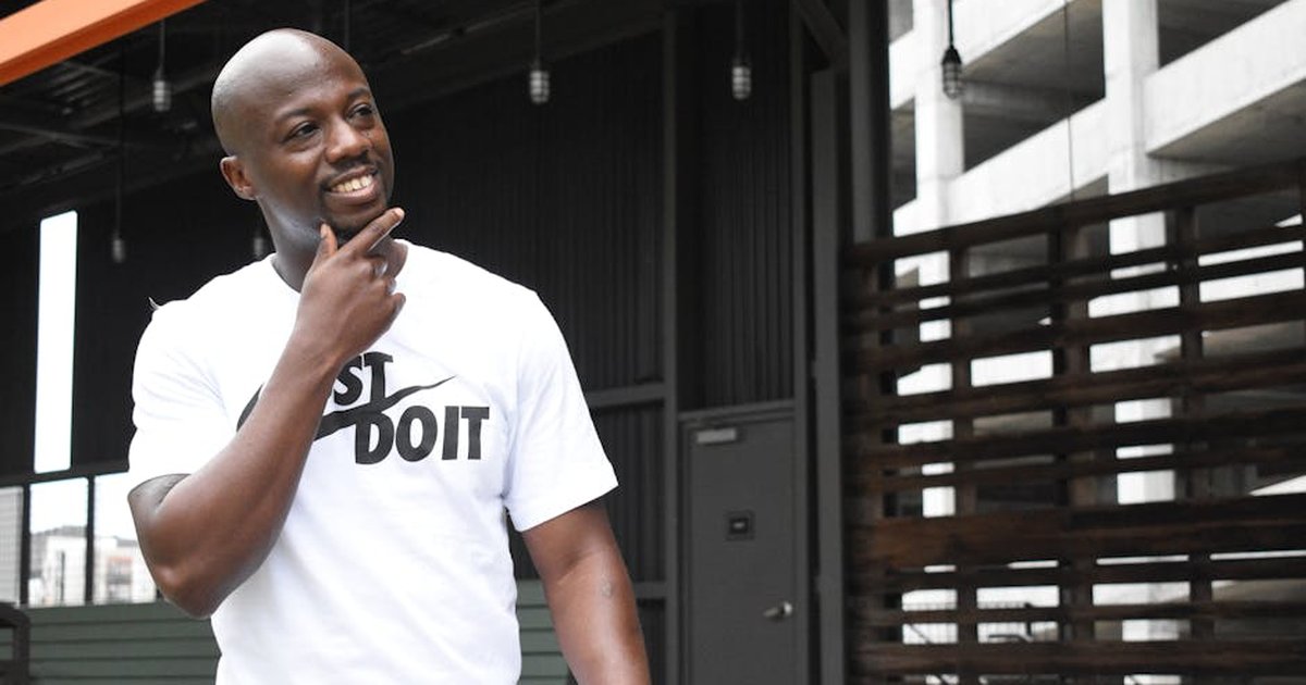 Smiling man in a white t-shirt stands confidently in an industrial outdoor setting.