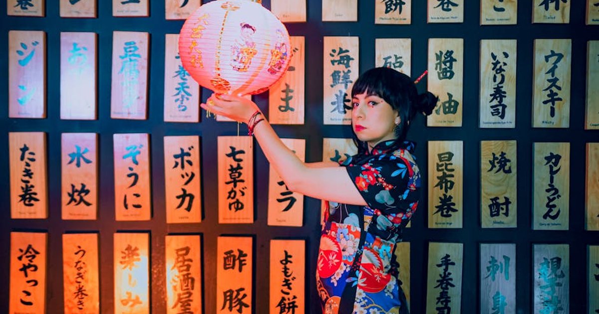 A woman in traditional attire holding a Japanese lantern against a backdrop of Japanese writings.