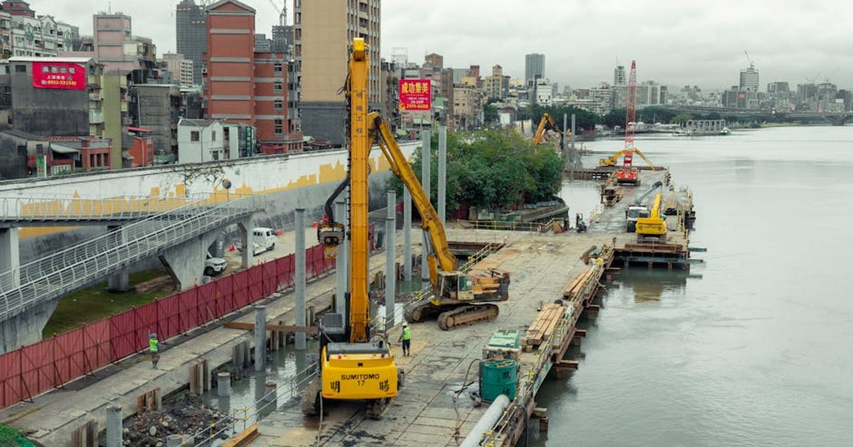 Industrial construction site by the river in Taipei with cranes and buildings in the background.