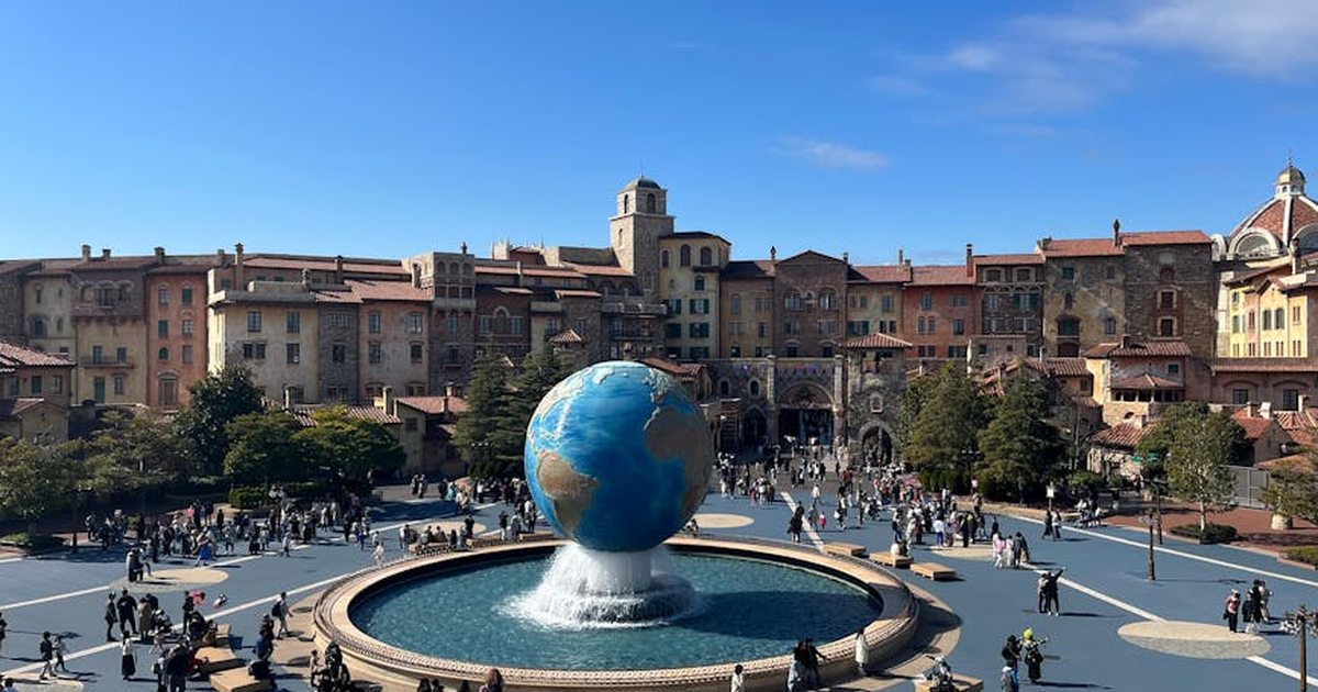 Wide view of a colorful piazza with a central globe fountain, bustling with people under a clear blue sky.