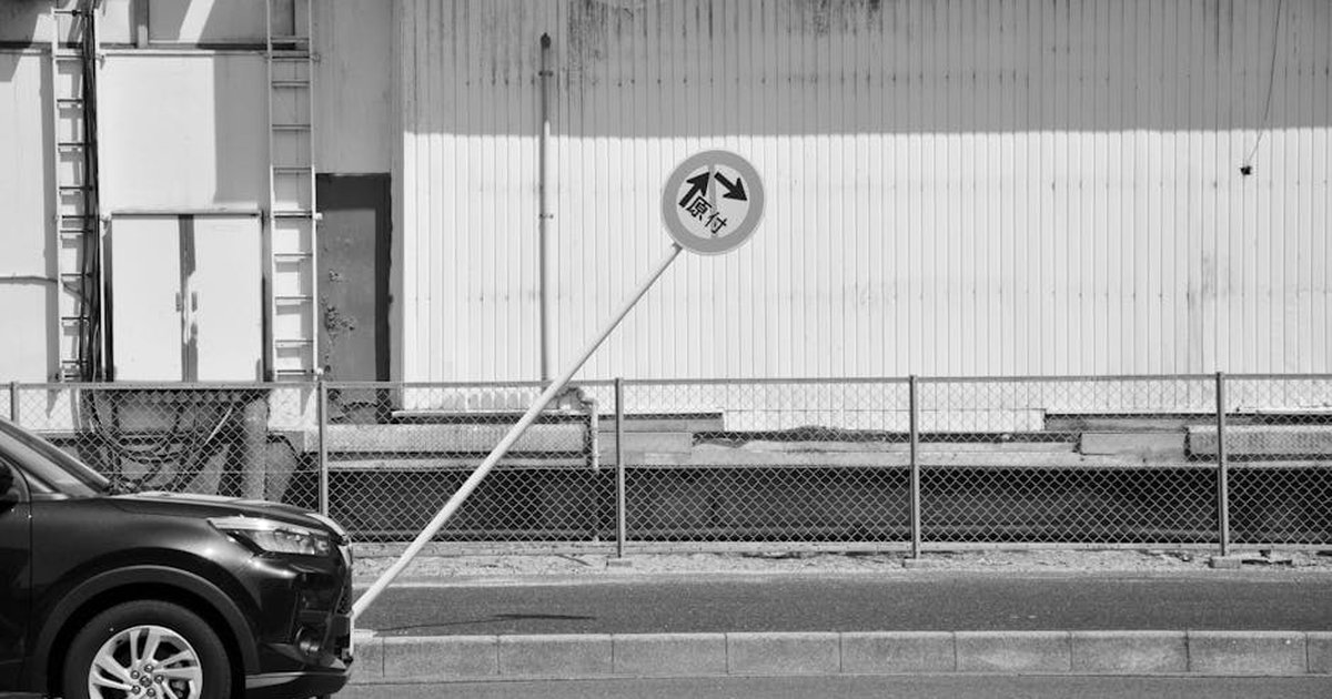 Black and white image of a leaning traffic sign beside a parked car and industrial wall.