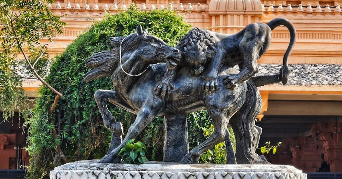 Bronze statue of a lion attacking a horse, set in a lush outdoor setting.