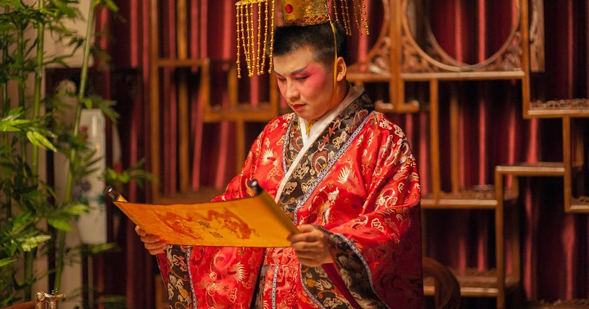 A person in traditional Chinese costume reading a scroll in a decorated room.