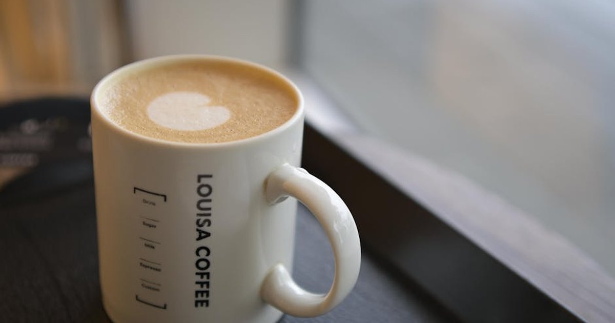 A hot latte with heart foam art in a Louisa Coffee mug on a wooden table by a window.