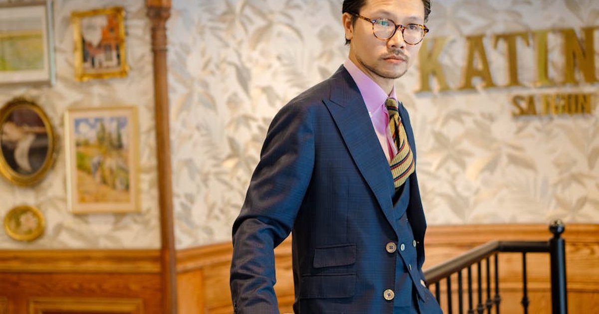 Stylish Asian man in elegant suit leaning indoors with a coffee cup, showcasing sophistication.