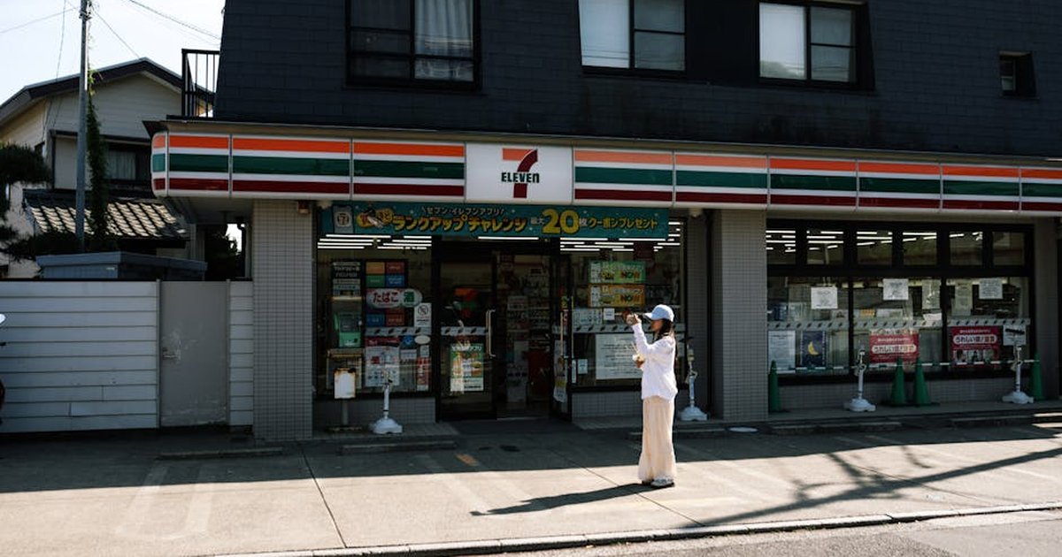A person taking a photo outside a 7-Eleven store on a sunny day with clear skies.