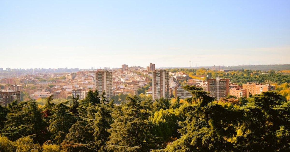 Panoramic cityscape view featuring lush green trees in the foreground and urban landscape in the background.