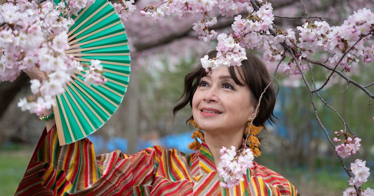 Woman in vibrant kimono holding fan beneath blossoming cherry trees, enjoying spring outdoors.