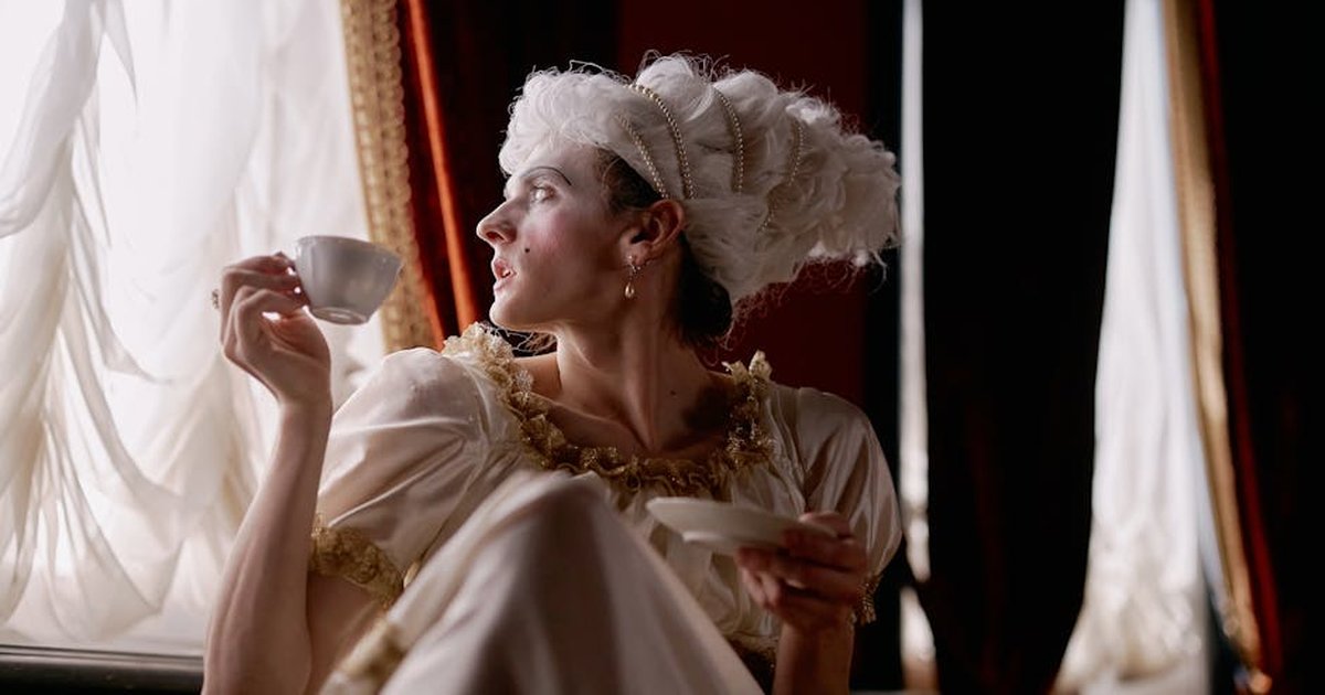A woman in historical costume enjoys tea by the window, exuding elegance and timelessness.