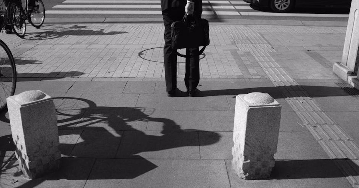 Black and white photo of a person standing on a city sidewalk, holding a bag, with bicycles and cars nearby.