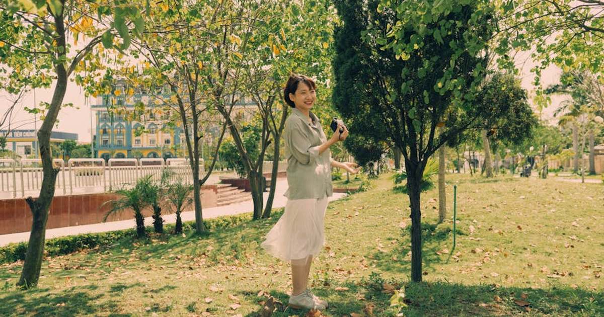 Asian woman smiling while holding a camera in a lush green park on a sunny day.