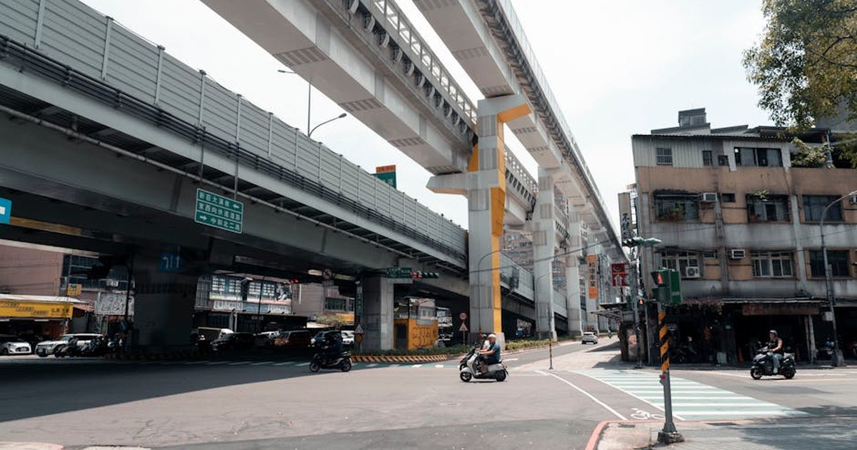 Motorbikes navigate an urban intersection beneath towering elevated train tracks in bustling New Taipei City.