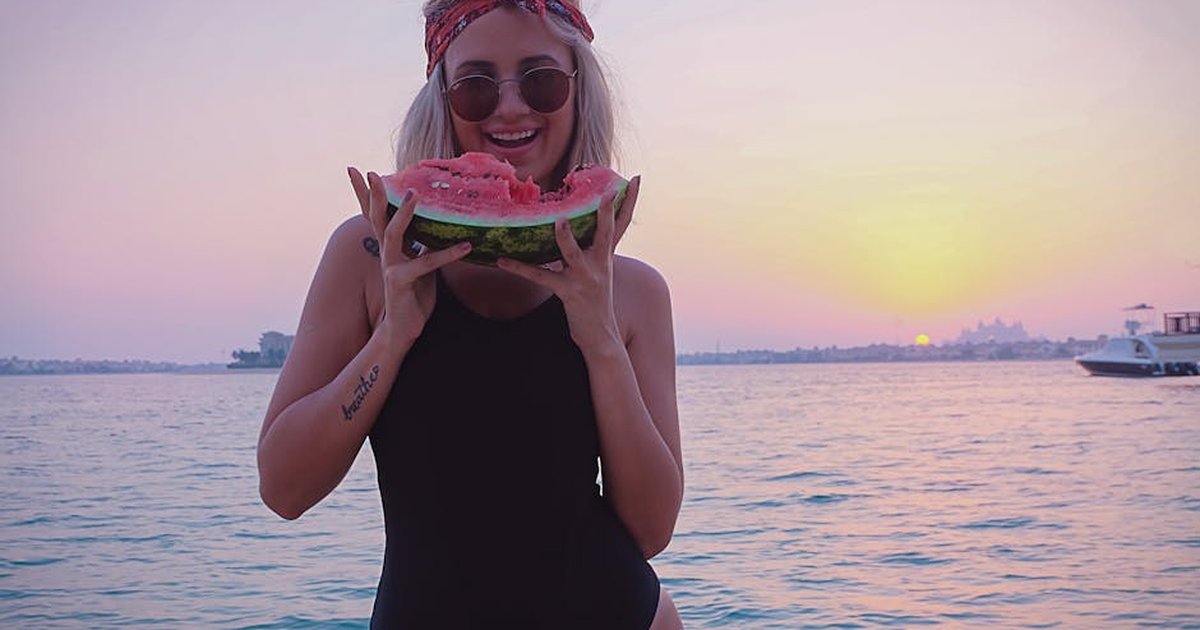 A woman in a bikini enjoys a slice of watermelon by the beach during a picturesque Dubai sunset.