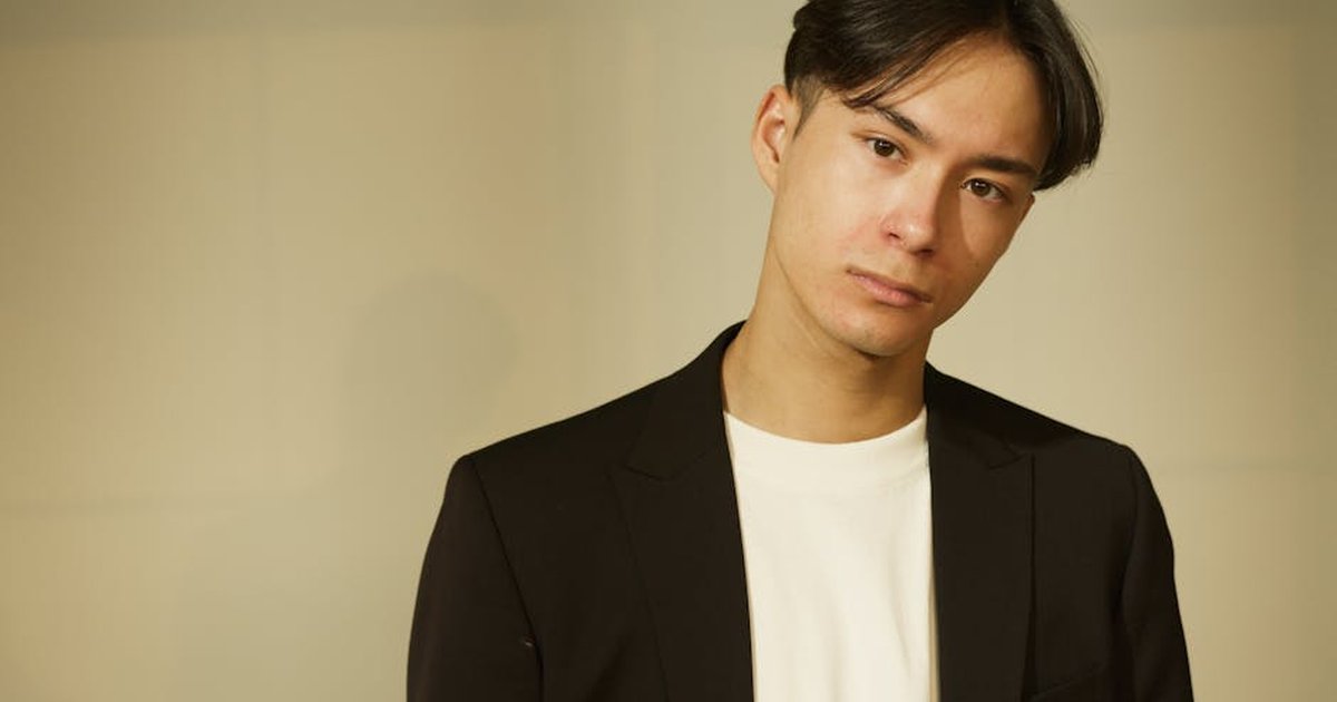 A young man in a black suit jacket, photographed in a studio setting with a neutral expression.