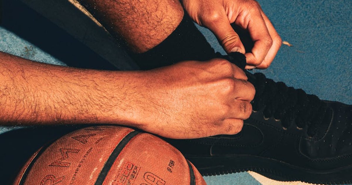 A person ties their basketball shoe on a sunlit outdoor court beside an official game ball.
