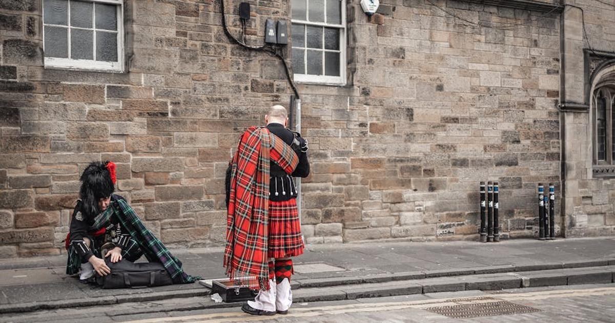 Two bagpipers in traditional Scottish kilts prepare instruments on a cobblestone street.