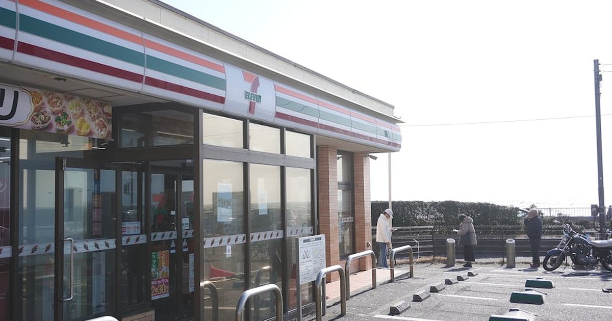 Exterior view of a 7-Eleven store with people, parking area, and clear skies.