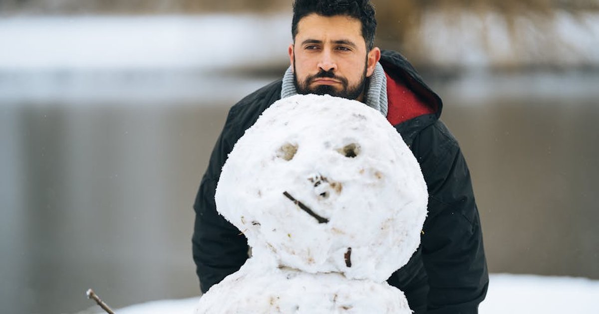 A bearded man poses with a snowman beside a lake during winter.