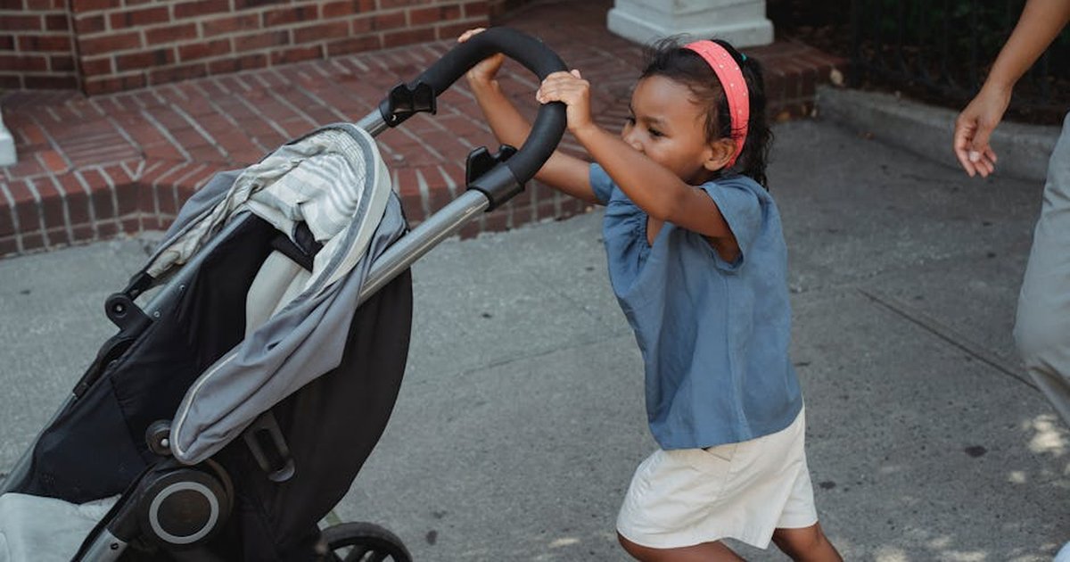 Side view of cheerful Asian preschool girl pushing baby stroller while walking on sidewalk near crop mother on street in city