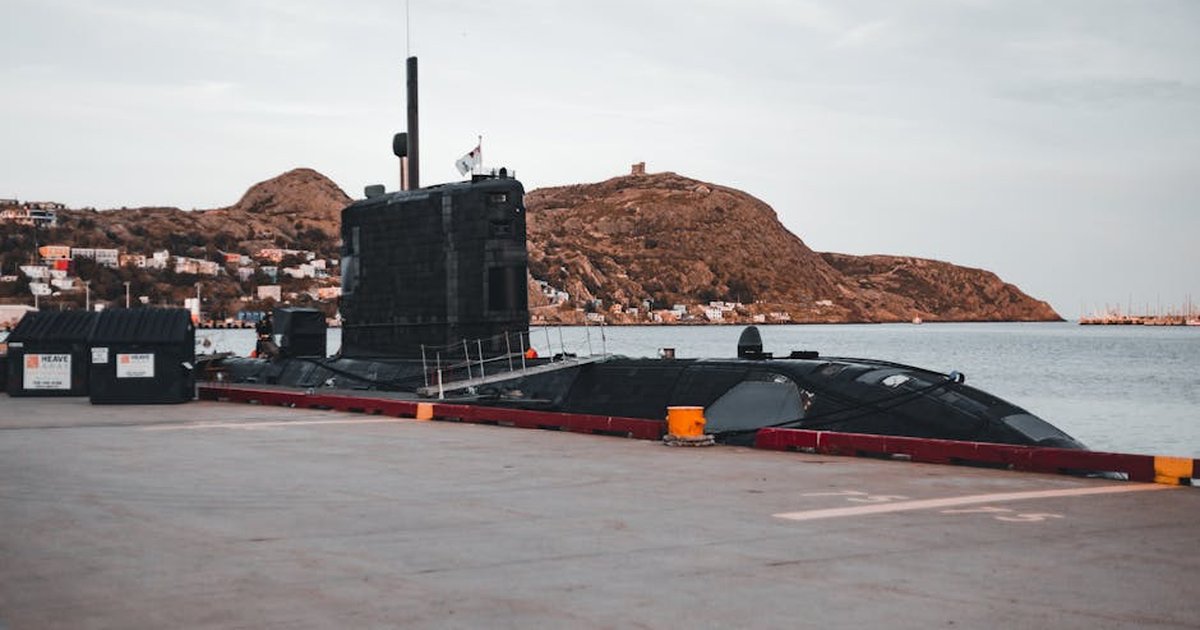 A submarine docked at an urban waterfront with scenic hills in the background.
