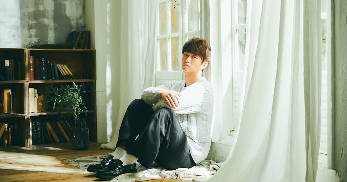 A fashionable man sits thoughtfully by a sunlit window in a cozy room.