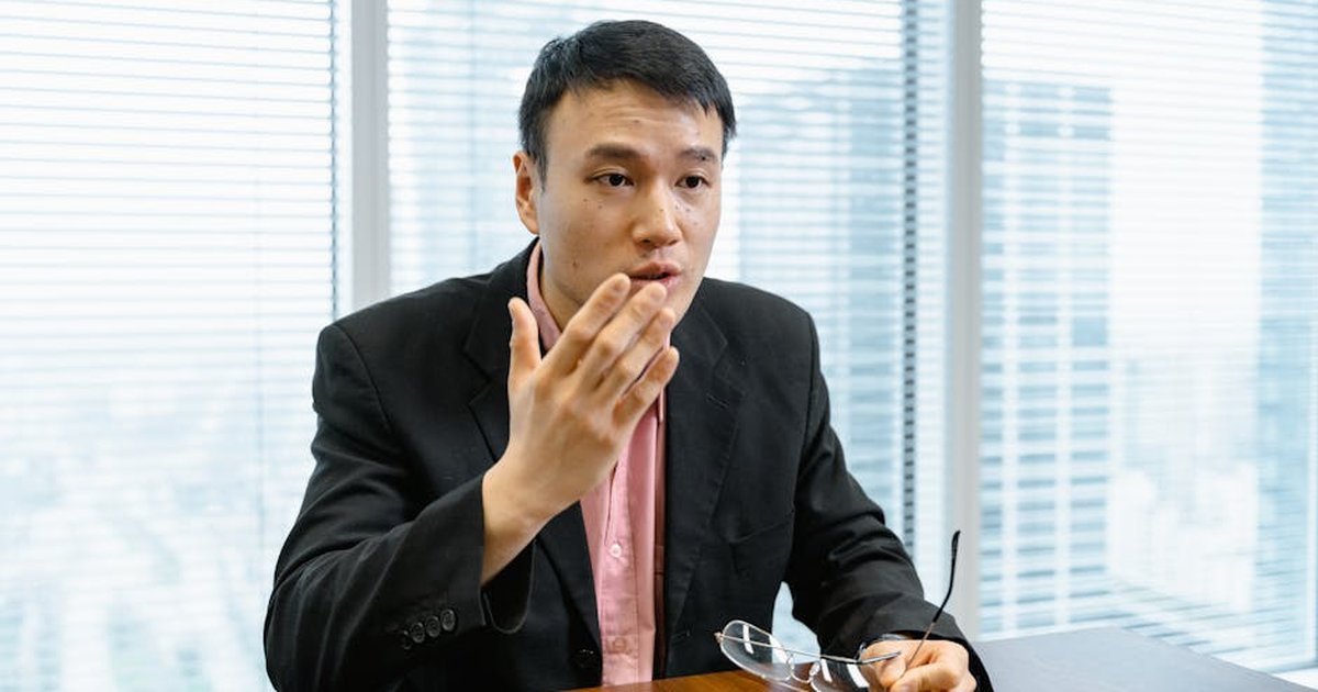 Asian man in black blazer speaking in an office with a city view behind.