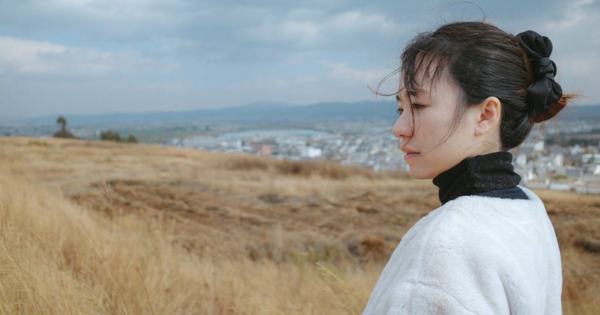 A thoughtful woman stands in a windswept field overlooking a town under cloudy skies.