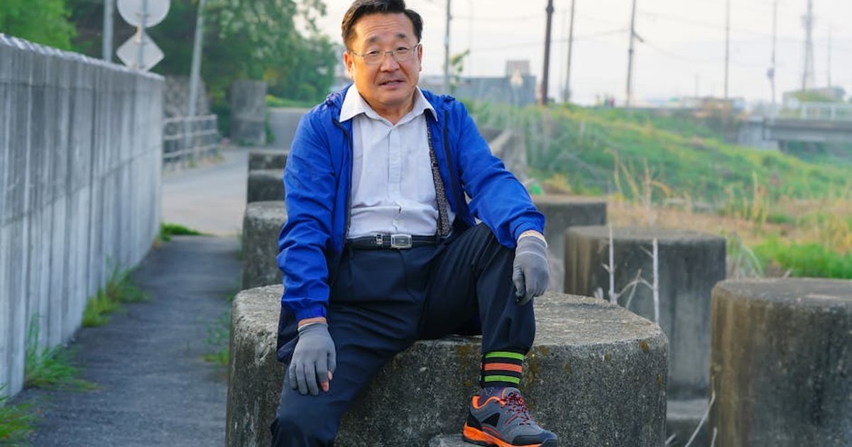 Older man with glasses sitting outside on concrete blocks in a casual pose.