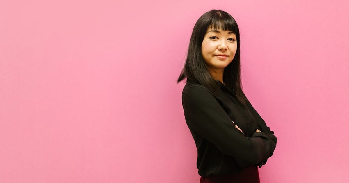 A confident woman in a black outfit standing against a pink background, arms crossed.
