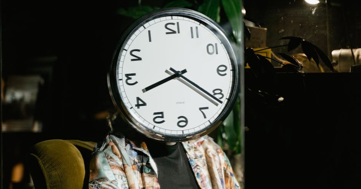 A person sits with a large wall clock covering their face, creating a surreal and artistic visual.