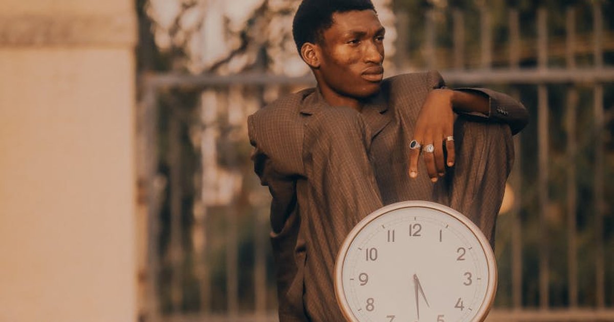 A contemplative man in a suit sits outdoors holding a large clock, embodying the passage of time.